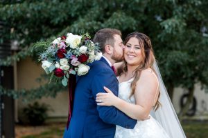 A young couple embrace at their wedding. The bride smiles for the camera, confident in her expertly styled hair and makeup.